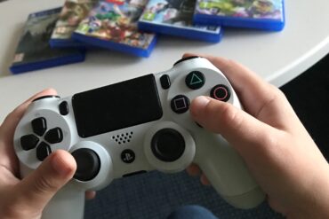 Närbild på barnhänder som håller i en PS4-kontroll. I bakgrunden ligger några olika tv-spel.